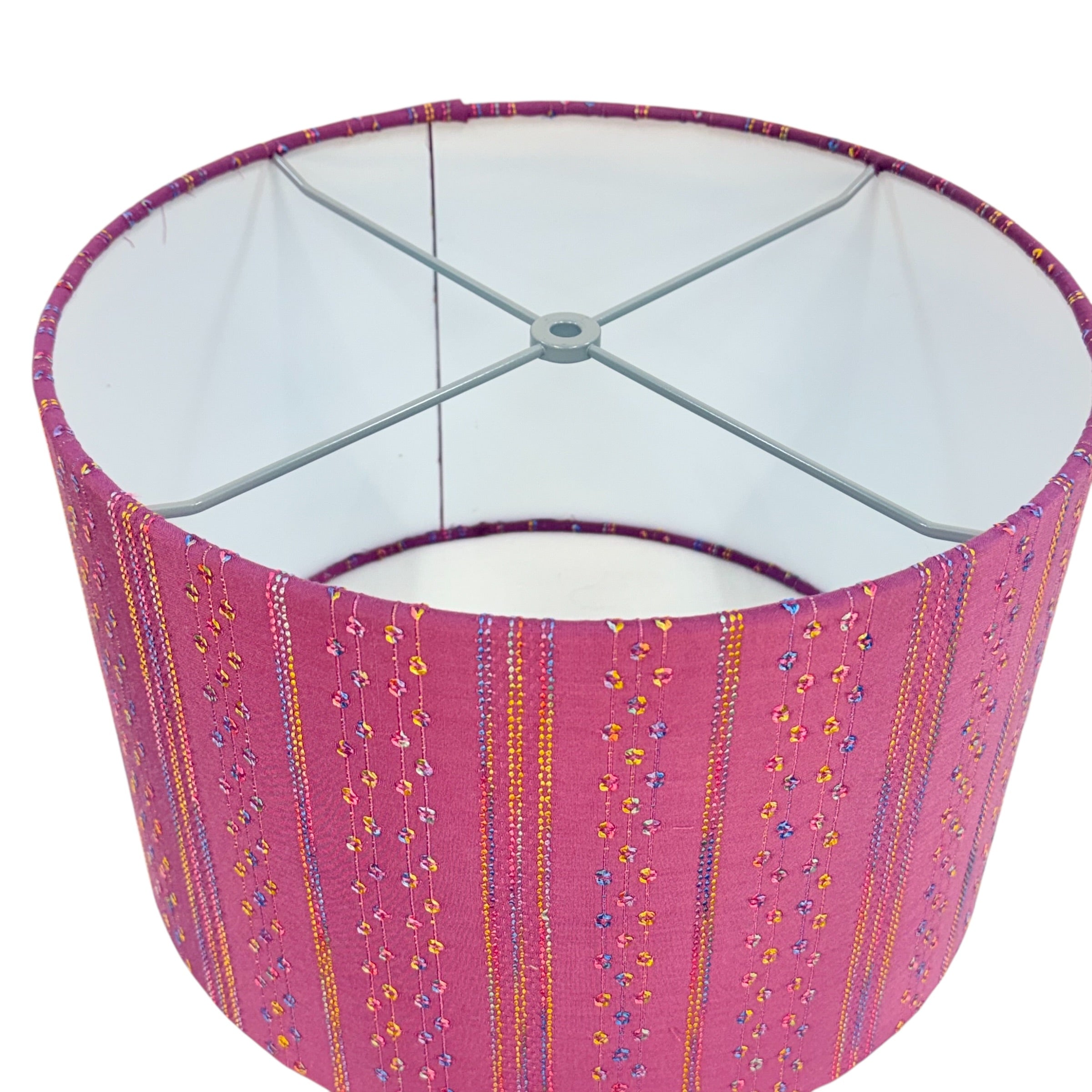 A cylindrical fabric lampshade with a vibrant pink base decorated with vertical stripes of gold, orange, and blue floral patterns, featuring a white interior and a gray metal spider support.