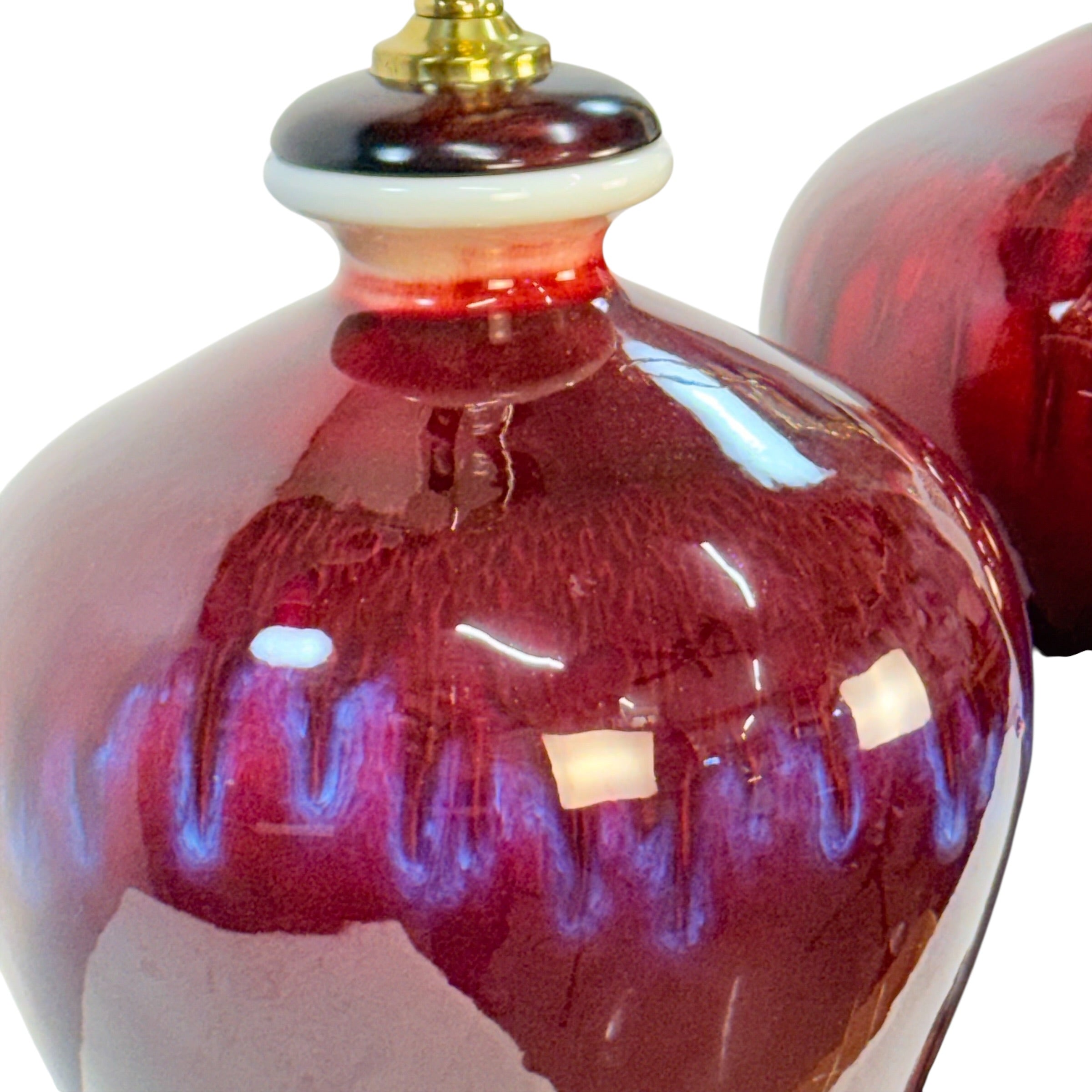 A close-up of a glossy ceramic vase or lidded container in deep burgundy red with blue iridescent drip-like glaze accents and a small gold-toned finial on a white background.
