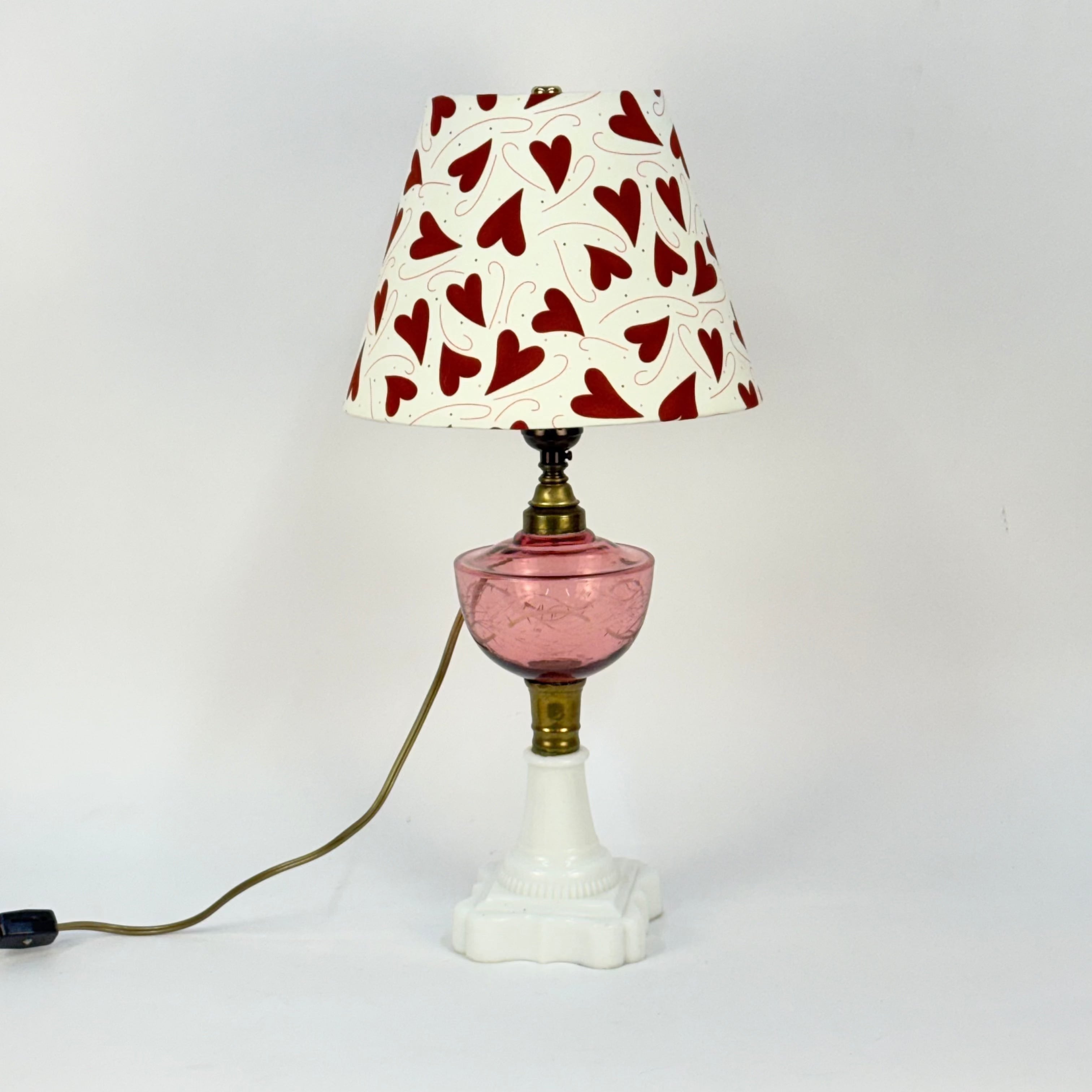 Lamp with a pink base, white shade with red heart pattern, and white stand on a light gray background