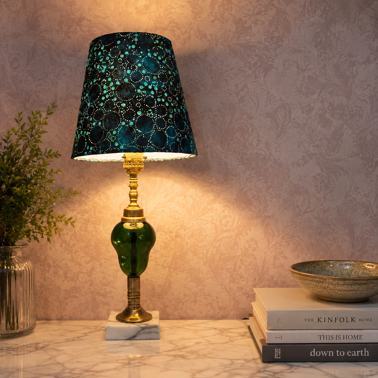 A decorative table lamp with a dark teal and black patterned shade, a green glass base, and gold-toned metal accents, placed on a marble surface against a patterned beige wall with books and a bowl nearby.