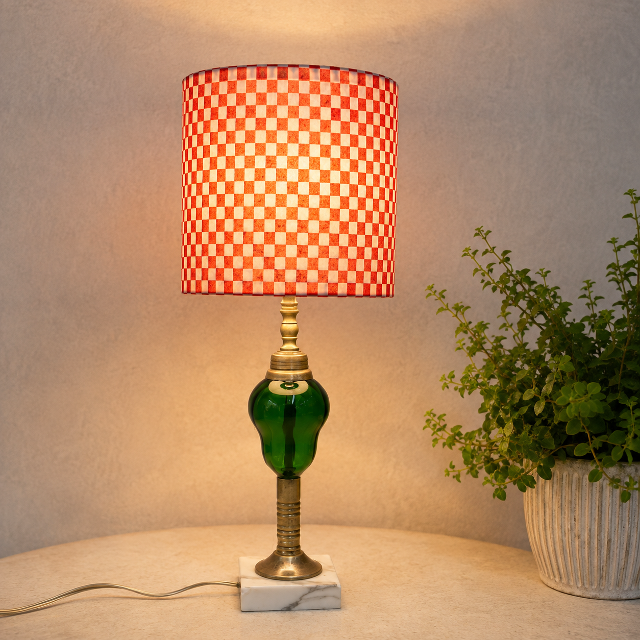 A table lamp with a red and white checkered shade, a green glass bulb, and a brass-toned base on a marble foot, shown lit on a tabletop next to a potted plant.