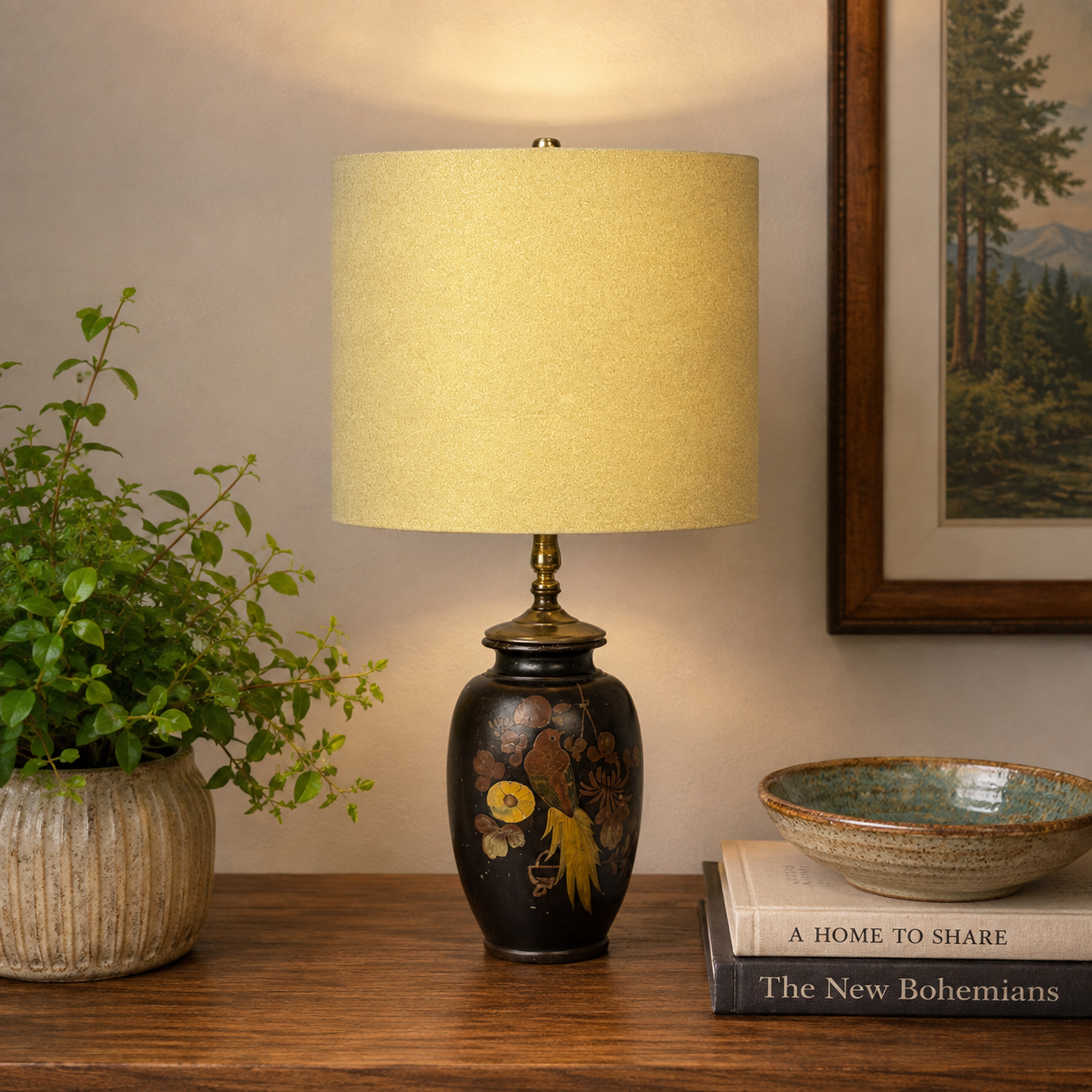 A decorative table lamp with a warm yellow fabric drum shade and a glossy black ceramic base decorated with gold and yellow floral motifs, styled on a wooden table next to a potted plant and books.