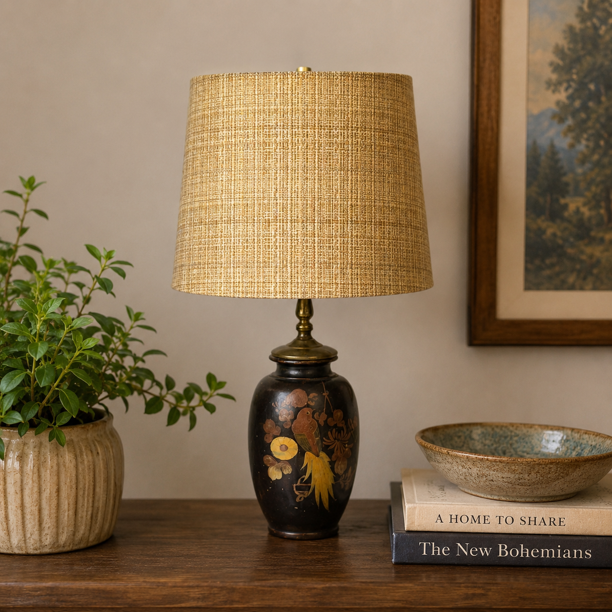 A table lamp with a textured beige woven drum shade and a glossy black ceramic base decorated with gold and brown floral motifs, styled on a wooden table next to a potted plant and books.