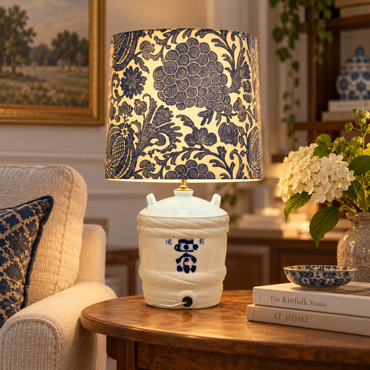 A decorative table lamp with a cream fabric drum shade featuring a dark blue floral and vine pattern, mounted on a white ceramic base with blue painted details, placed on a wooden table in a cozy living room setting.