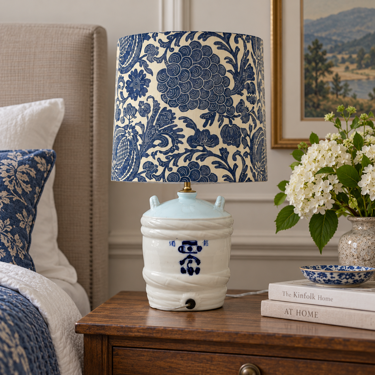 A table lamp with a blue and cream floral patterned drum shade and a white ceramic base decorated with blue motifs, styled on a wooden nightstand in a bedroom setting.