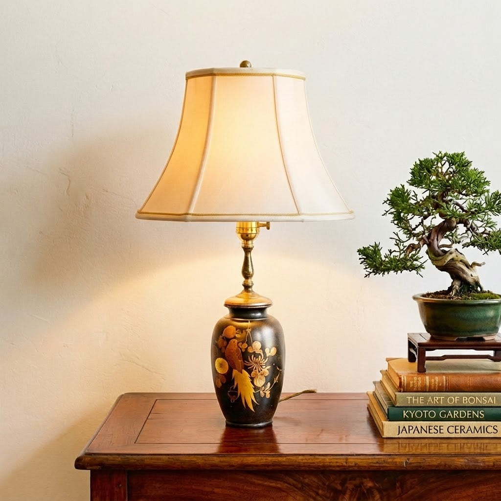 A decorative table lamp with a warm beige fabric shade and a glossy black ceramic base featuring gold and yellow floral motifs, styled on a wooden side table next to a small potted bonsai tree and books.