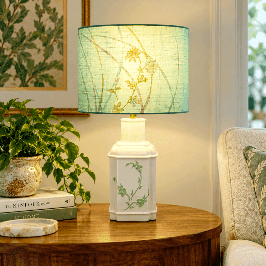 A table lamp with a white ceramic base decorated with green floral vines and a light green drum shade featuring a yellow wildflower print, styled on a wooden side table in a cozy living room setting.