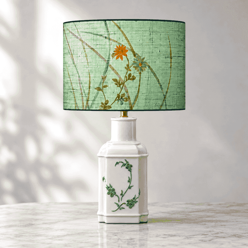 A table lamp with a white ceramic bottle-shaped base decorated with green botanical illustrations, topped with a sage green drum shade featuring a floral print of an orange flower and green stems.