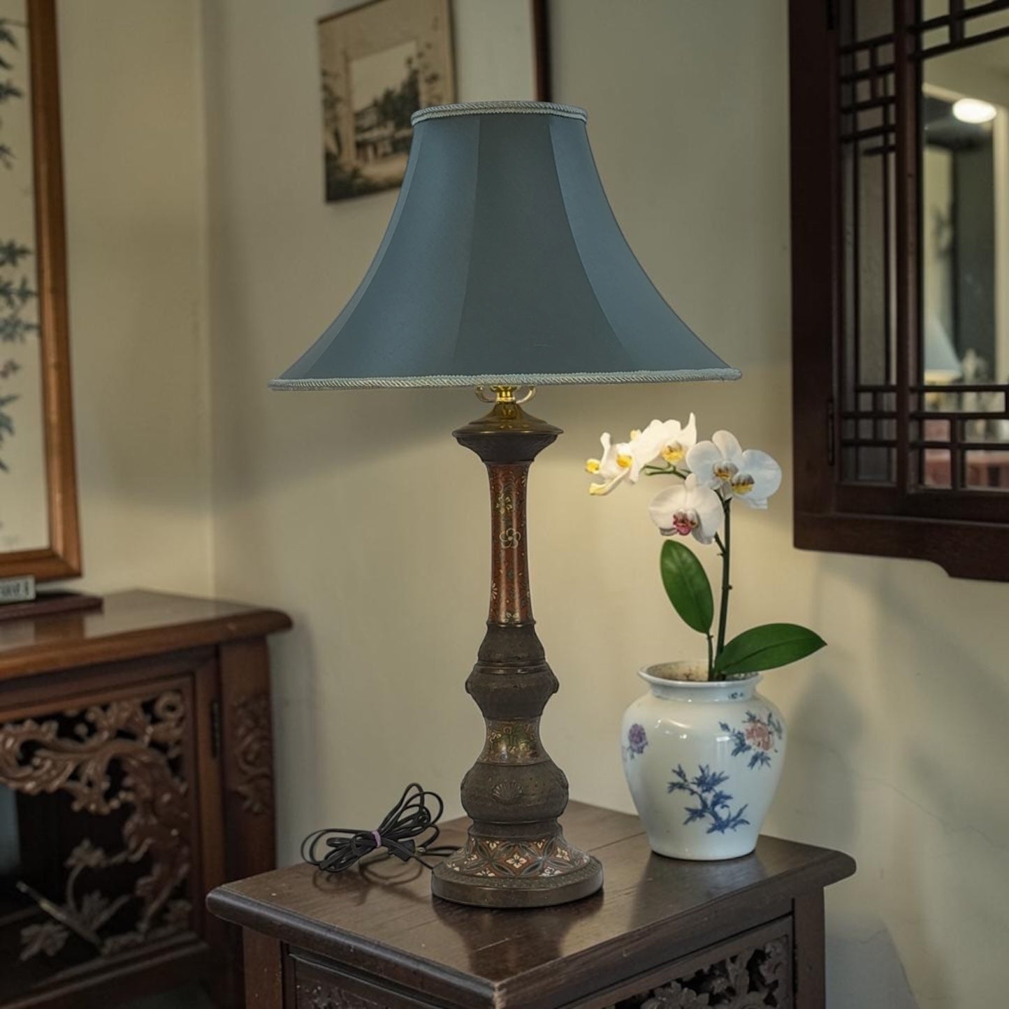 Decorative table lamp with a blue shade on a wooden side table next to a vase with flowers.