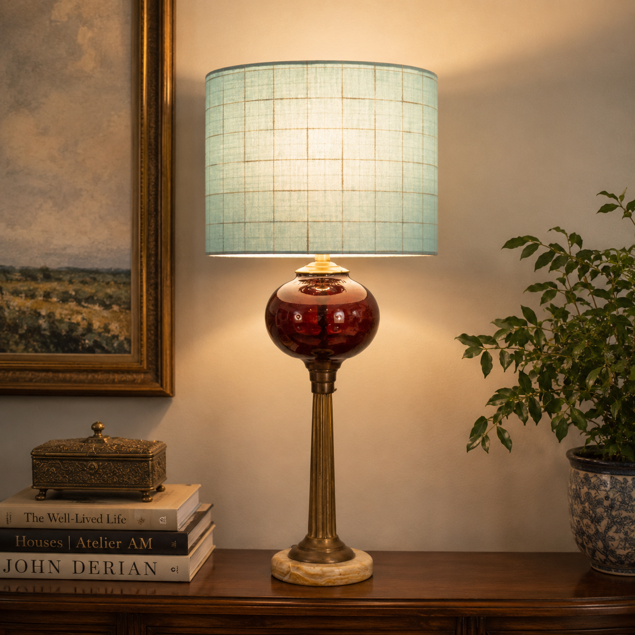 A lit table lamp with a light blue grid-patterned drum shade, a glossy dark red glass globe base, and a brass-toned fluted stem and marble-like foot, shown on a wooden console beside a framed painting, books, and a potted plant.