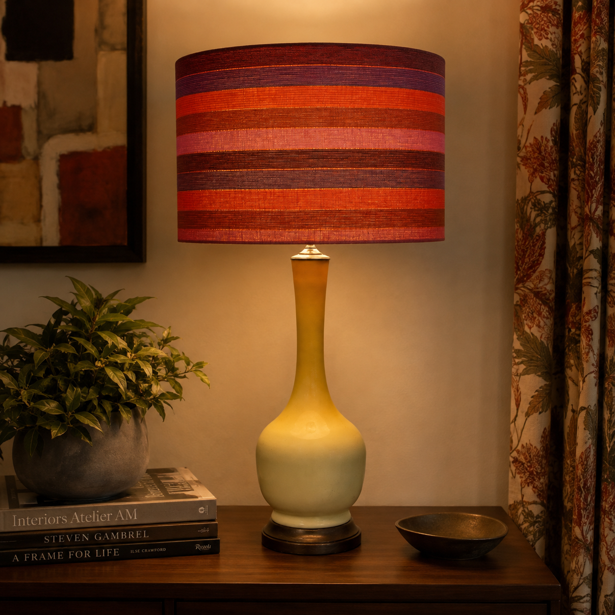 A table lamp with a striped fabric shade in red, orange, and purple tones, paired with a glossy ceramic base that fades from pale yellow to cream, shown lit on a wooden console table in a warmly lit room.