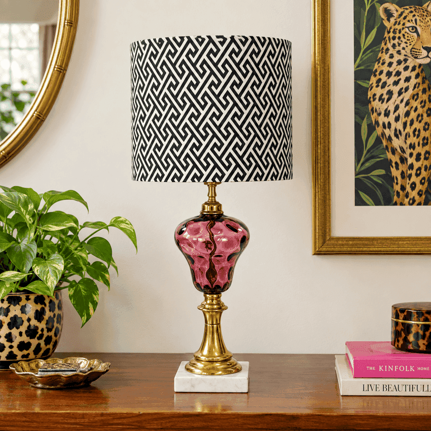 An amethyst thumbprint glass table lamp with a black and white geometric patterned drum shade, a glossy pink glass bulb, and a brass-finished base on a white marble pedestal, displayed on a wooden console table in a styled room setting.