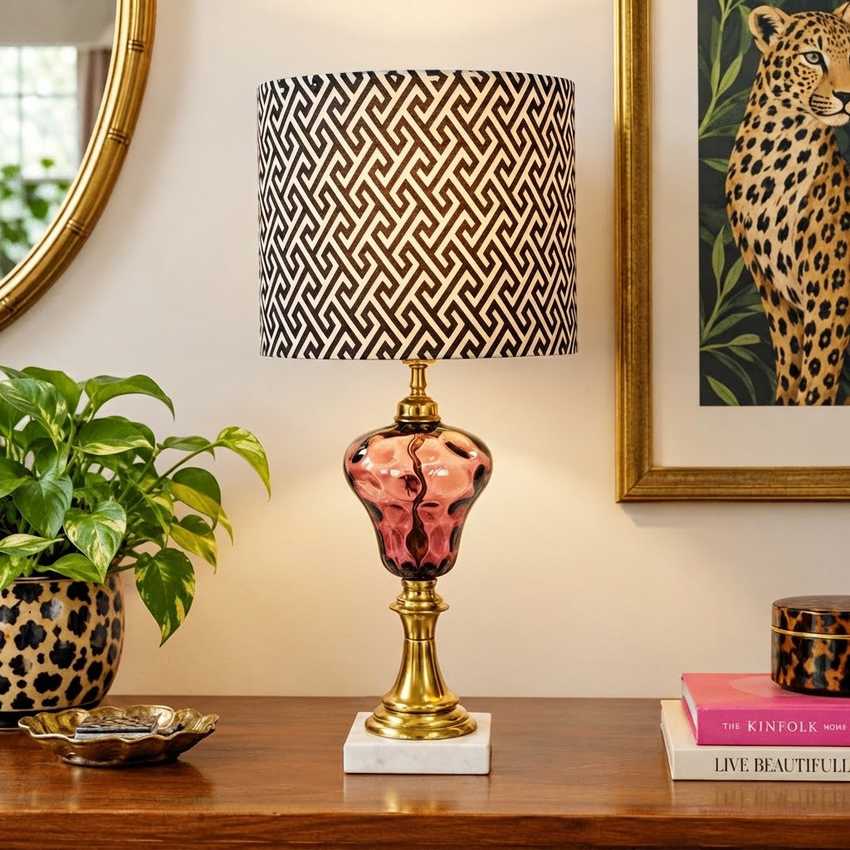 A decorative table lamp with a black and beige geometric patterned drum shade, a glossy amethyst thumbprint glass body, and a brass-finished base on a white marble pedestal, displayed on a wooden console table in a styled room setting.