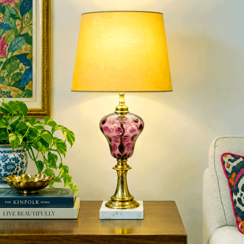 A decorative table lamp with a warm yellow fabric shade, a gold-toned brass lower body and marble base, and a amethyst thumbprint glass bulb body, displayed on a wooden side table in a styled room setting.