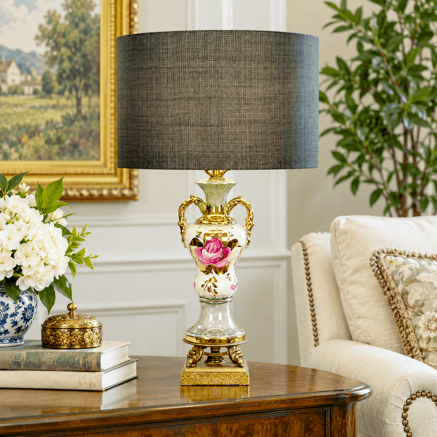 An ornate table lamp with a black silk  textured drum shade and a porcelain urn base featuring gold accents and a pink rose, shown on a wooden sideboard in a styled living room setting.