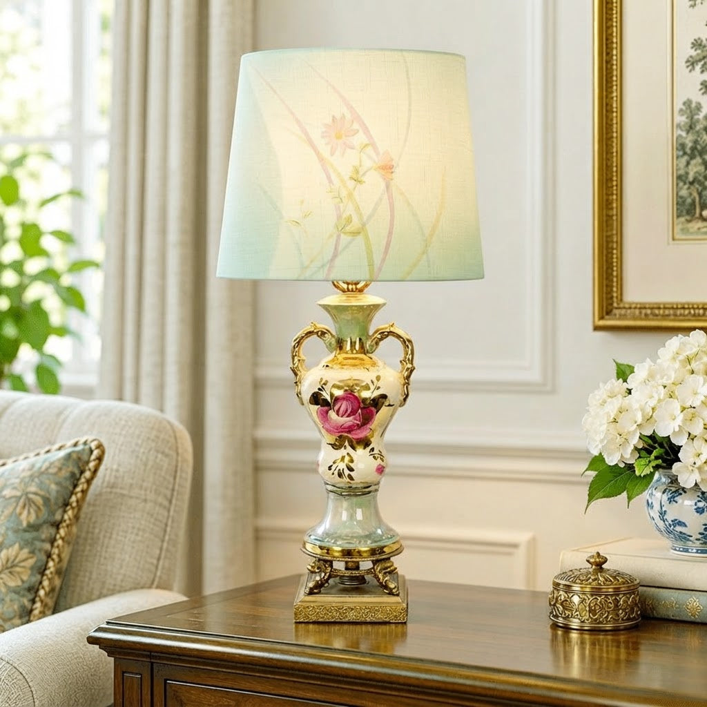 An ornate table lamp with a pale blue-green and gold ceramic vase body, featuring a pink rose motif, set on a wooden console table in a bright living room next to a sofa and flowers.