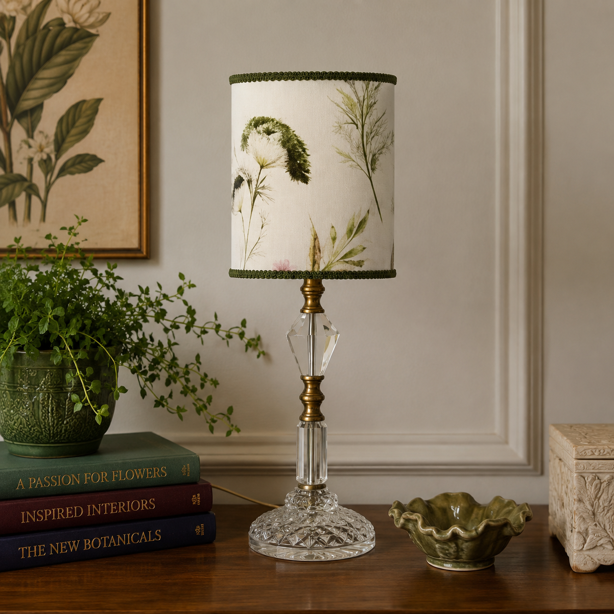A table lamp with a cream fabric shade printed with botanical illustrations, a clear faceted crystal base, and a brass-toned stem, styled on a wooden table with books, a plant, and a small bowl in a room setting.