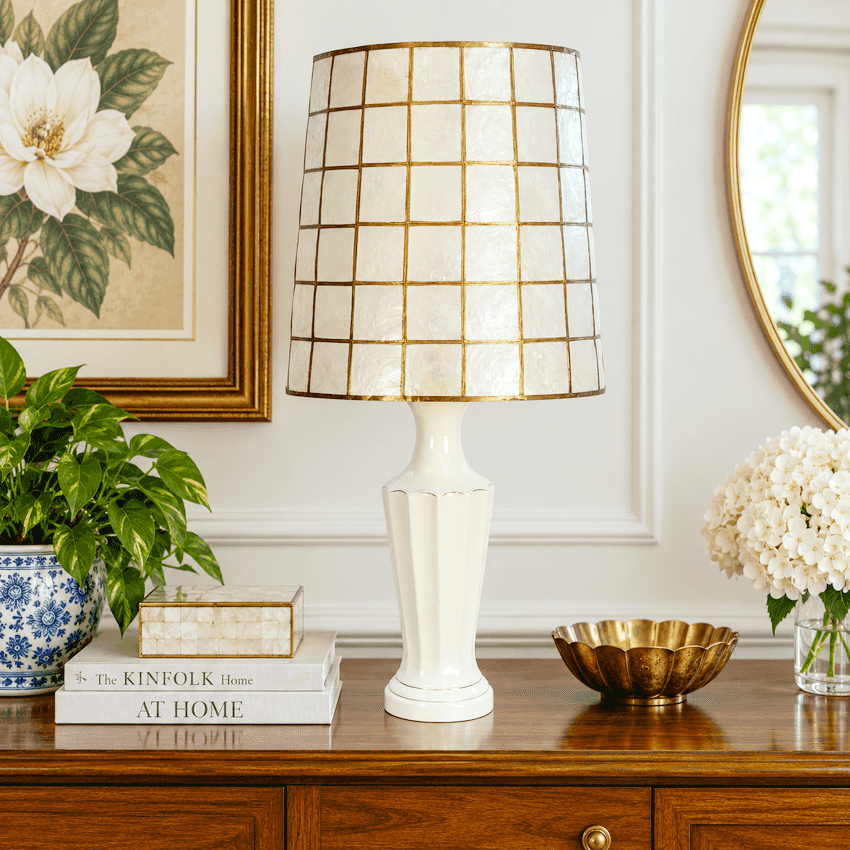 A table lamp with a white ceramic fluted base and a cream-colored shade featuring a gold grid pattern, lit up on a wooden console table in a warm, elegant room setting.