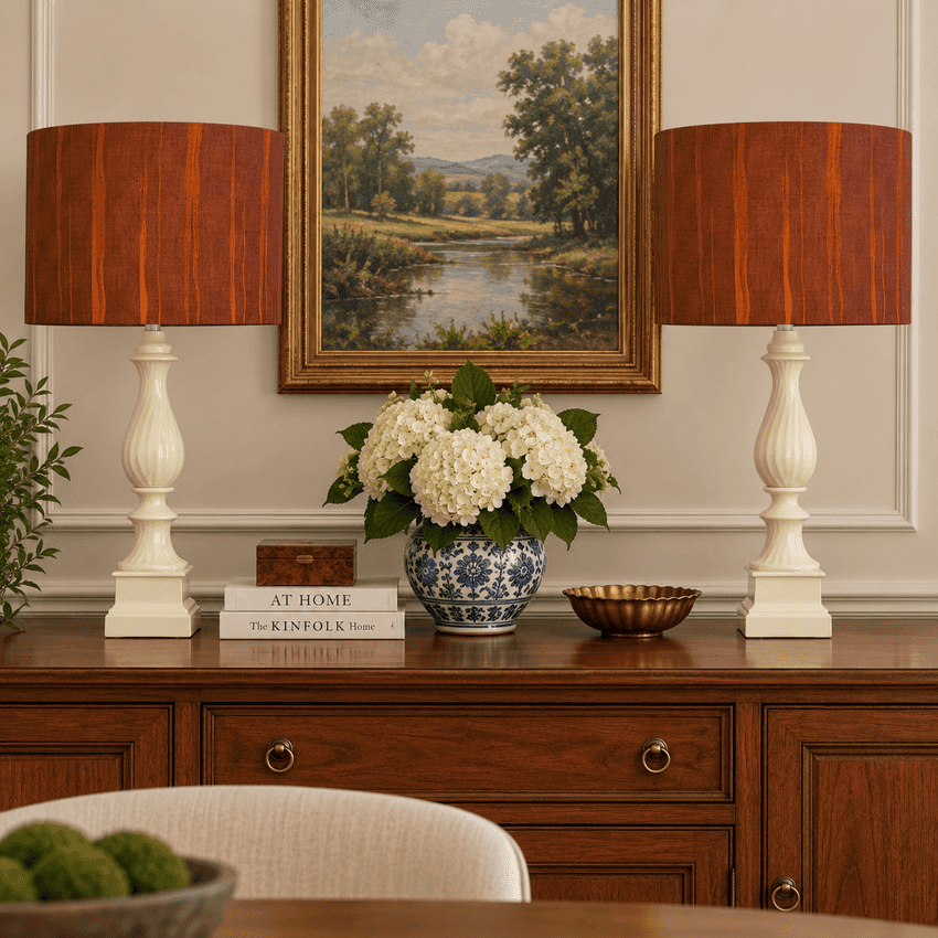 Two matching table lamps with reddish-brown wood-look drum shades and cream pedestal bases sit on either side of a floral arrangement on a wooden console table, with a framed landscape painting above.