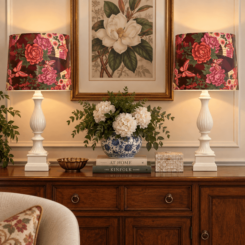A styled interior scene showing two matching table lamps with floral lampshades on a wooden console table, flanking a blue-and-white ceramic vase with white flowers. A framed floral print hangs above, and part of a chair is visible in the lower left.