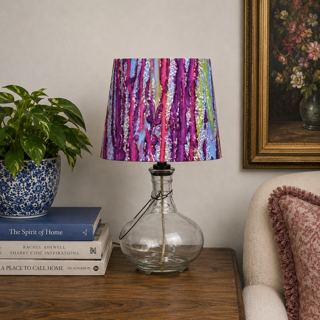 A table lamp with a colorful abstract tree-bark patterned shade in purple, pink, blue, and green, mounted on a clear glass bottle base, styled on a wooden table next to a potted plant and a chair.
