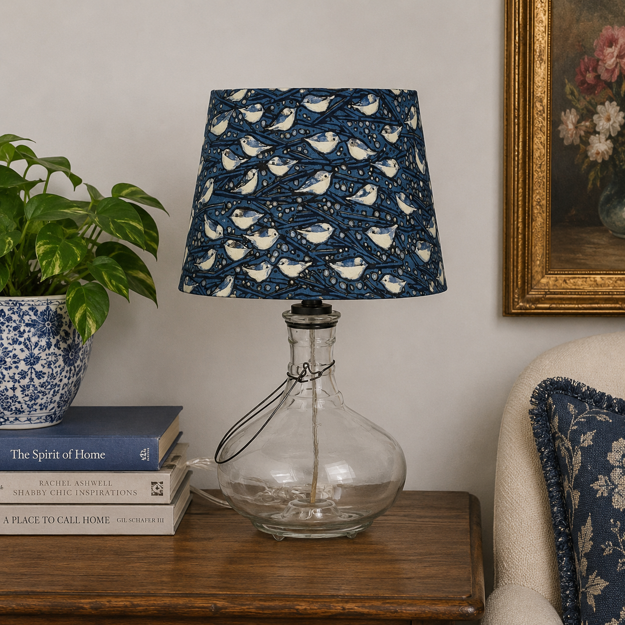 A table lamp with a navy blue fabric shade featuring a repeating pattern of white birds, set on a dark wooden side table next to a potted plant and books.