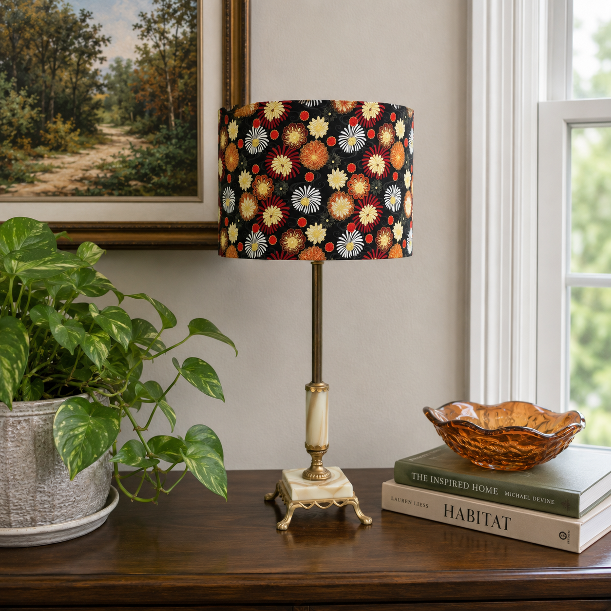 A table lamp with a black drum shade covered in a colorful floral and daisy-like pattern, standing on a dark wooden console table in a styled room. The lamp has a brass-toned stem and base with a cream-colored cylindrical accent.