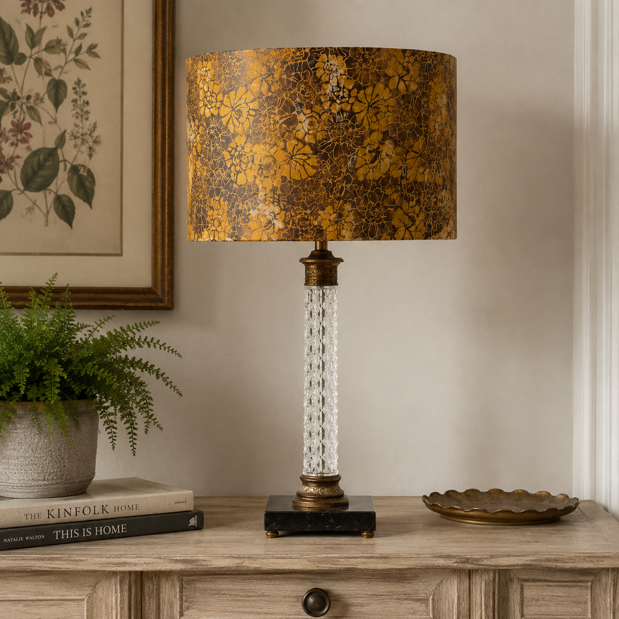 A table lamp with a textured gold and brown floral drum shade, a clear faceted crystal column stem, and an antique bronze base, styled on a wooden console table next to a potted fern and a decorative tray.