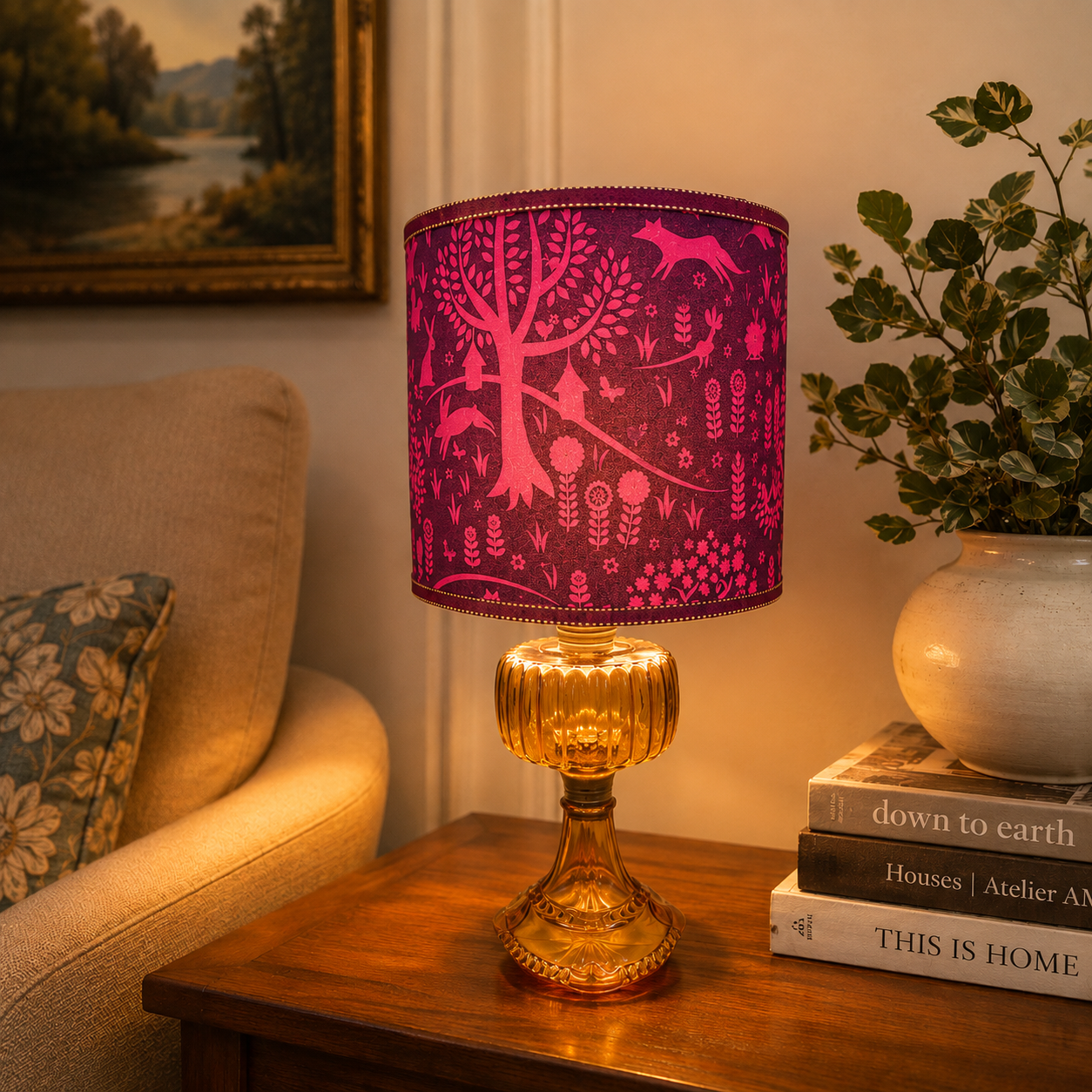 A table lamp with a vibrant magenta pink fabric drum shade featuring a white silhouette pattern of a tree, birds, and foliage, mounted on a ribbed amber glass pedestal base. The lamp is styled on a wooden side table in a cozy living room setting next to a sofa and a potted plant.