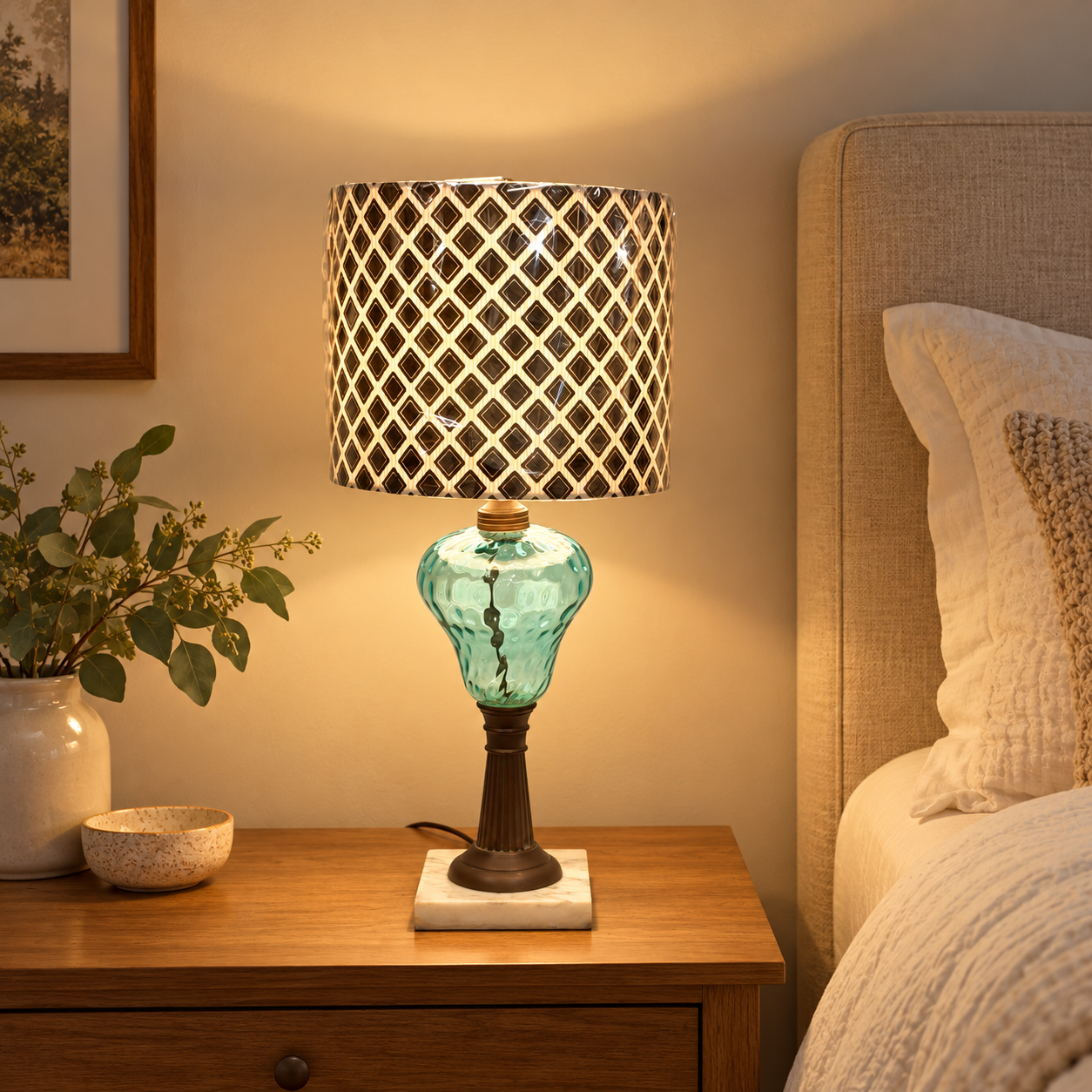 A table lamp with a turquoise glass base and a black-and-gold diamond-patterned drum shade, lit on a wooden bedside table next to a bed.