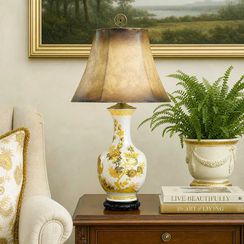 An ornate table lamp with a white ceramic vase body decorated with gold floral patterns and a textured bronze-gold conical shade, displayed on a wooden side table in a styled living room setting next to a potted fern.