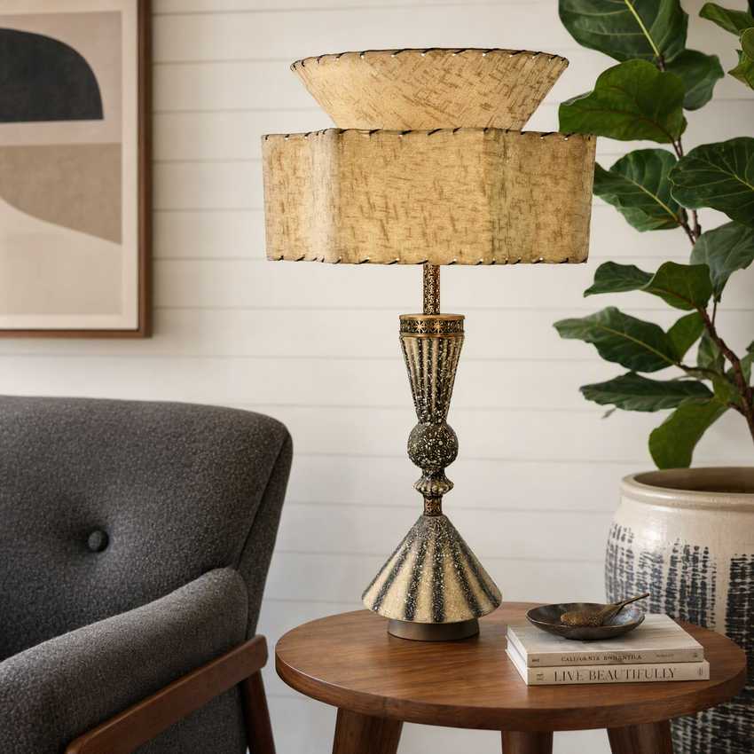 A decorative table lamp with a textured beige woven drum shade and a distressed gold and bronze metal base, styled on a wooden side table in a living room setting.
