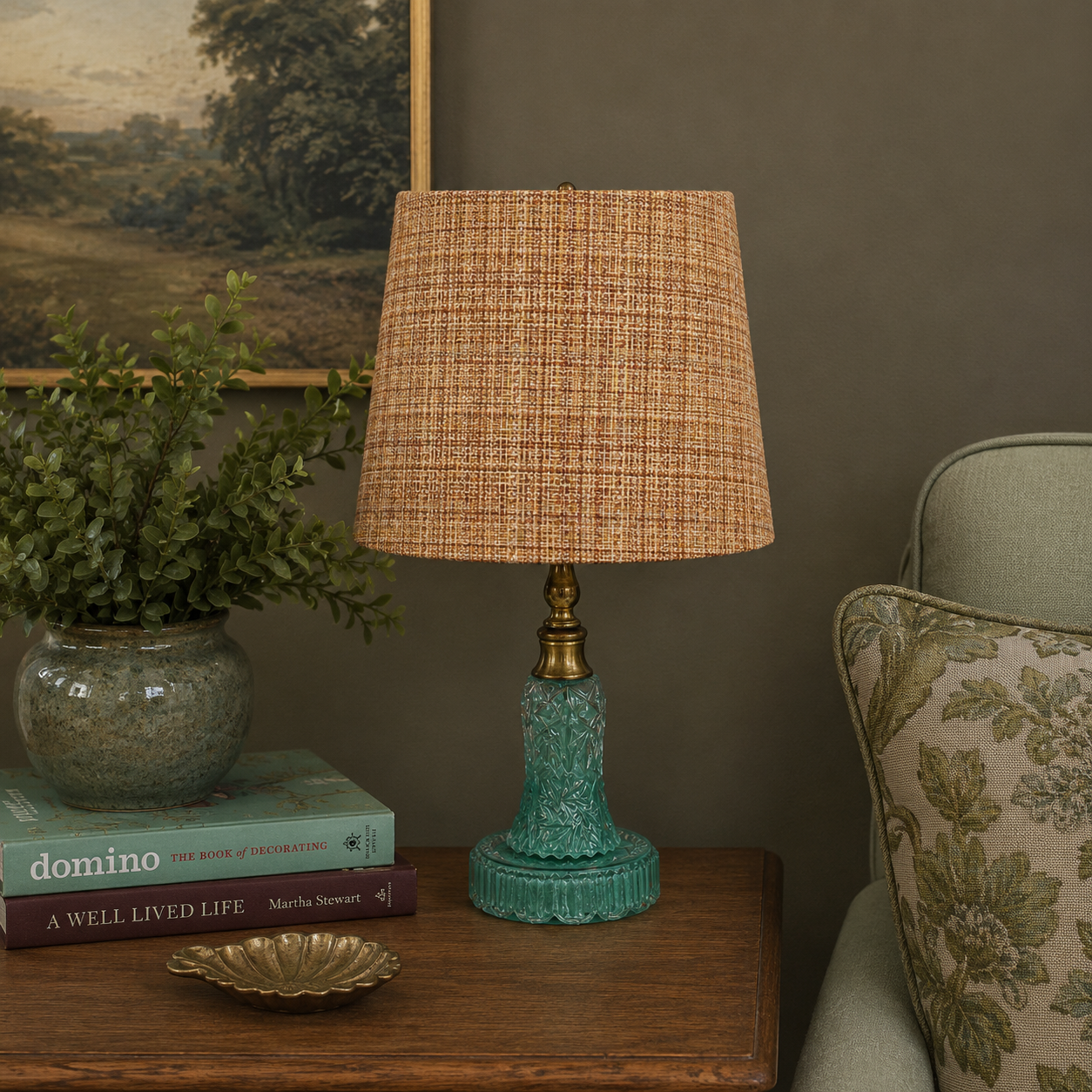 A table lamp with a textured tan woven shade and a turquoise glass base sits on a wooden side table in a styled room setting next to a chair and a potted plant.