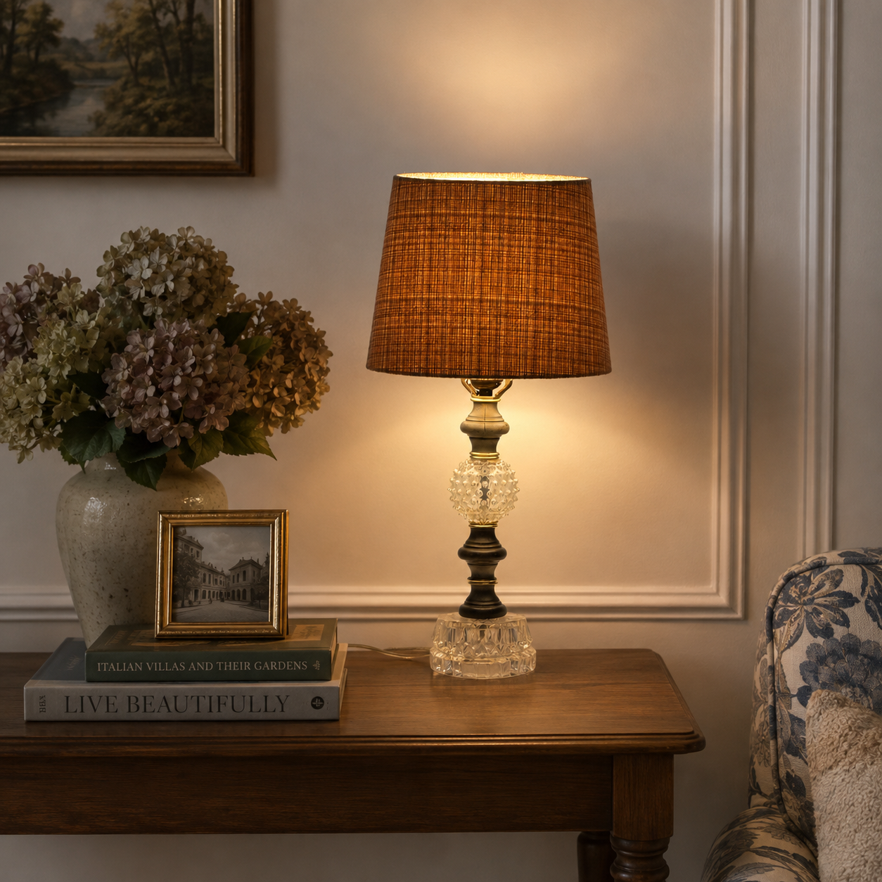 A classic table lamp with a textured brown fabric drum shade, a clear crystal prismatic base, and an antique bronze and gold-toned column, illuminated on a wooden console table in a cozy room setting.