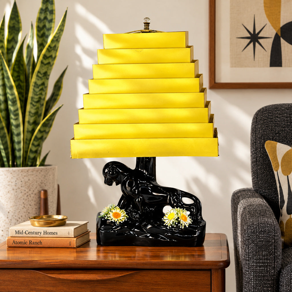 A decorative table lamp with a bright yellow tiered shade and a glossy black sculpted base featuring a reclining figure and small white flowers, shown on a wooden side table in a styled room with a plant, books, and chair.