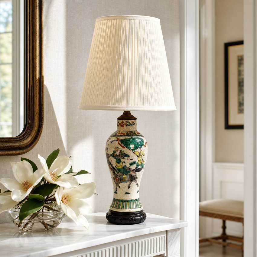A decorative table lamp with a pleated cream fabric shade and a ceramic base featuring a multicolor landscape scene, styled on a white console table next to white flowers in a bright room.