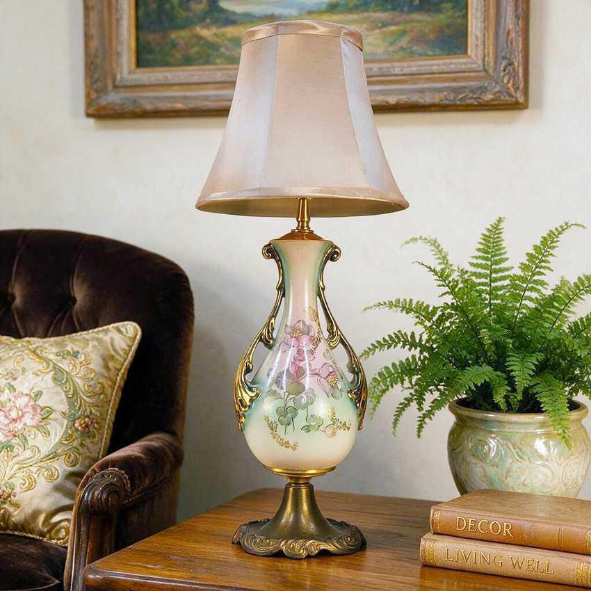 An ornate table lamp with a cream fabric shade and a porcelain base decorated with pink floral motifs and gold accents, styled on a wooden table next to a potted fern and books.