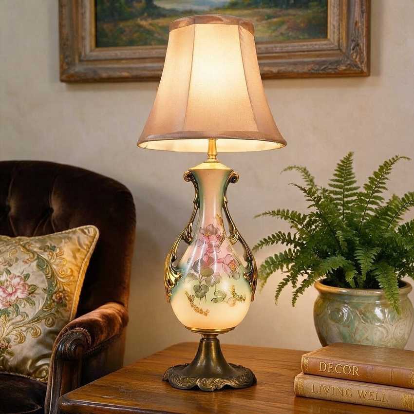 A lit table lamp with a beige fabric shade and an ornate ceramic base featuring a floral design, styled on a wooden table next to a chair, a potted fern, and books in a cozy room setting.