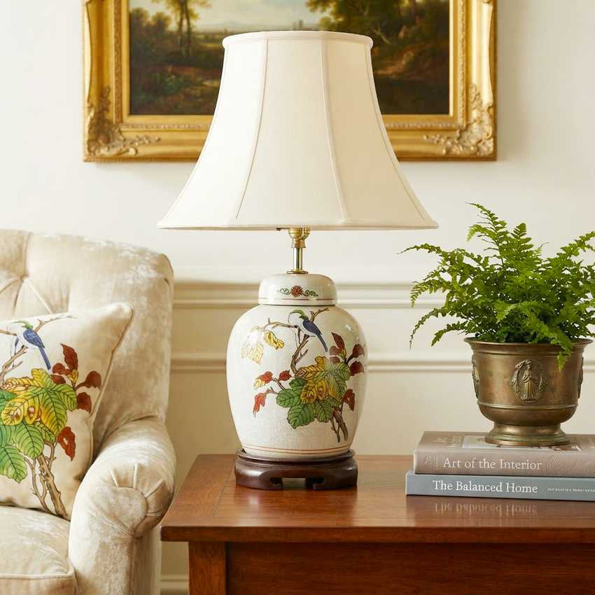 A ceramic table lamp with a cream pleated shade and a white jar-shaped base decorated with colorful birds and foliage, set on a wooden side table in a styled room next to an armchair and a potted fern.