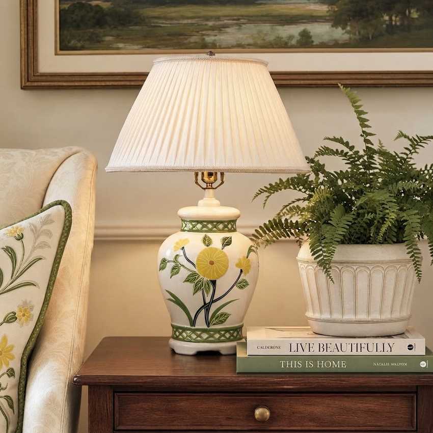 A decorative ceramic table lamp with a pleated cream shade and a white base decorated with yellow flowers and green leaves, styled on a wooden side table next to a potted fern and books.