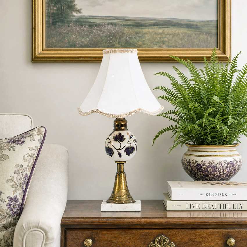A vintage-style table lamp with a white fabric shade, a white ceramic body decorated with purple floral motifs, and a gold-toned metal base, styled on a wooden side table next to a potted fern and a sofa.