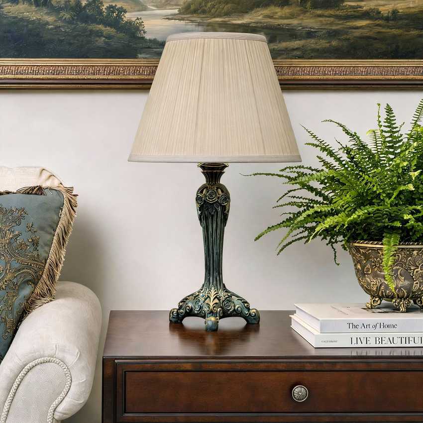 An ornate table lamp with a pleated beige fabric shade and a dark teal-green carved metal base sits on a dark wood side table in a styled living room setting.