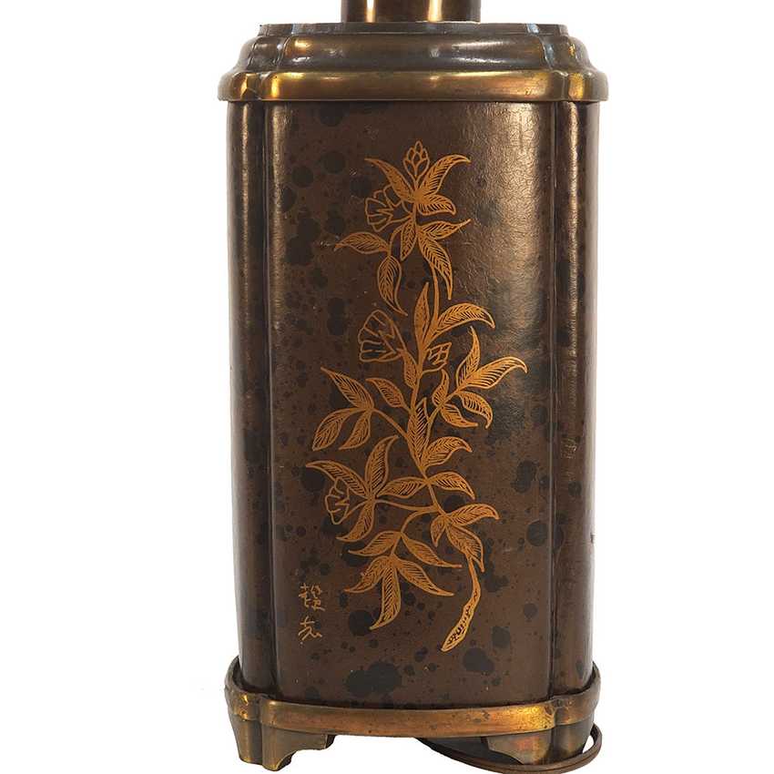 A vintage-style cylindrical metal container or lantern with a dark bronze-brown body, mottled dark spots, and gold-toned floral engravings on the front, shown against a plain white background.
