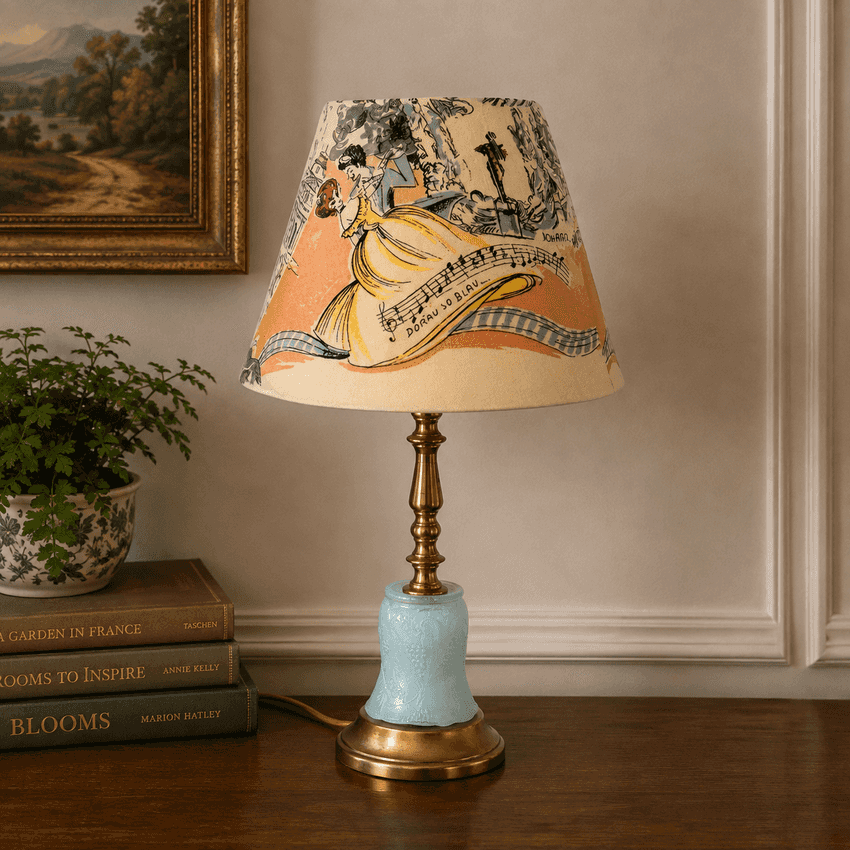 A vintage-style table lamp with a beige lampshade printed with a yellow dress and musical notes, a light blue crackled glass base, and a brass stem, set on a wooden table in a decorated room.