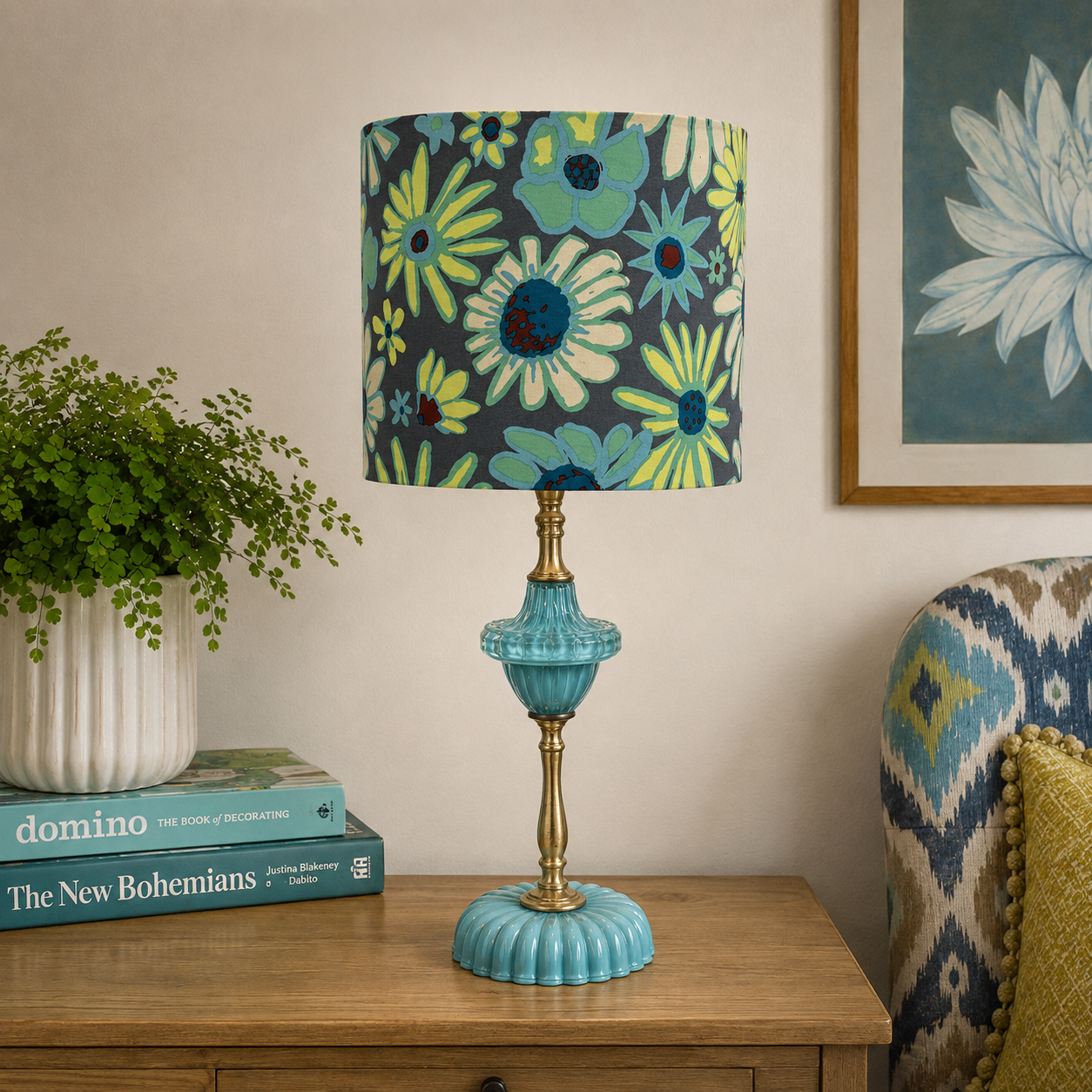 A decorative table lamp with a dark navy floral drum shade and a turquoise glass pedestal base sits on a wooden side table in a styled room setting.