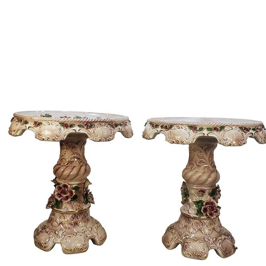 A pair of ornate vintage-style pedestal tables with scalloped cream-colored tops and bases, decorated with embossed floral details and small rose accents, shown against a plain white background.