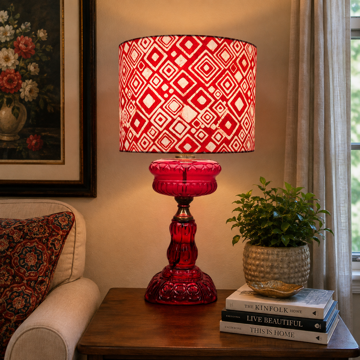 A red and white patterned table lamp with a faceted red glass base, styled on a wooden side table next to a potted plant and books in a cozy room setting.