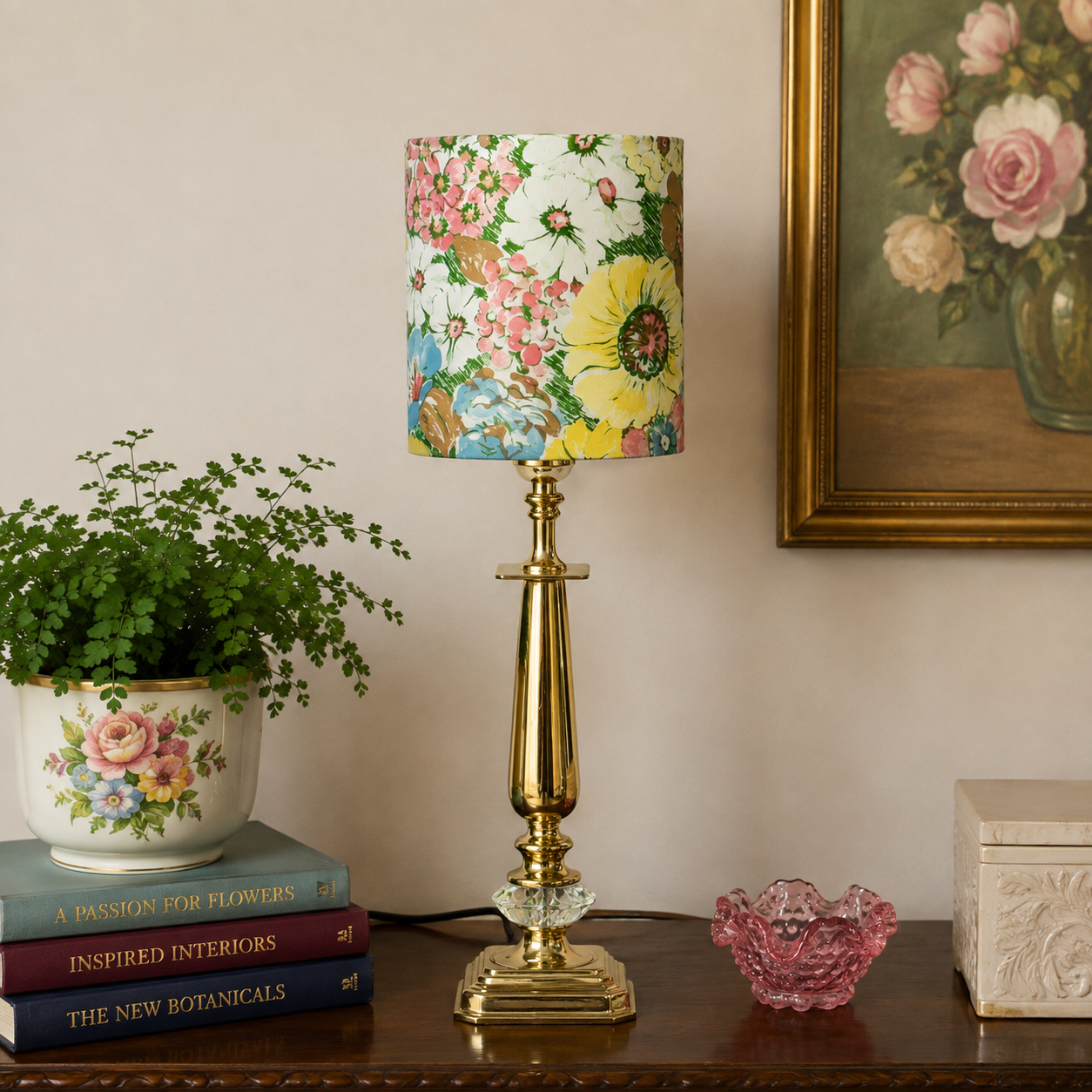 A brass table lamp with a colorful floral-patterned drum shade stands on a dark wooden table in a styled room setting, next to a potted plant, books, a pink glass bowl, and a framed floral painting.