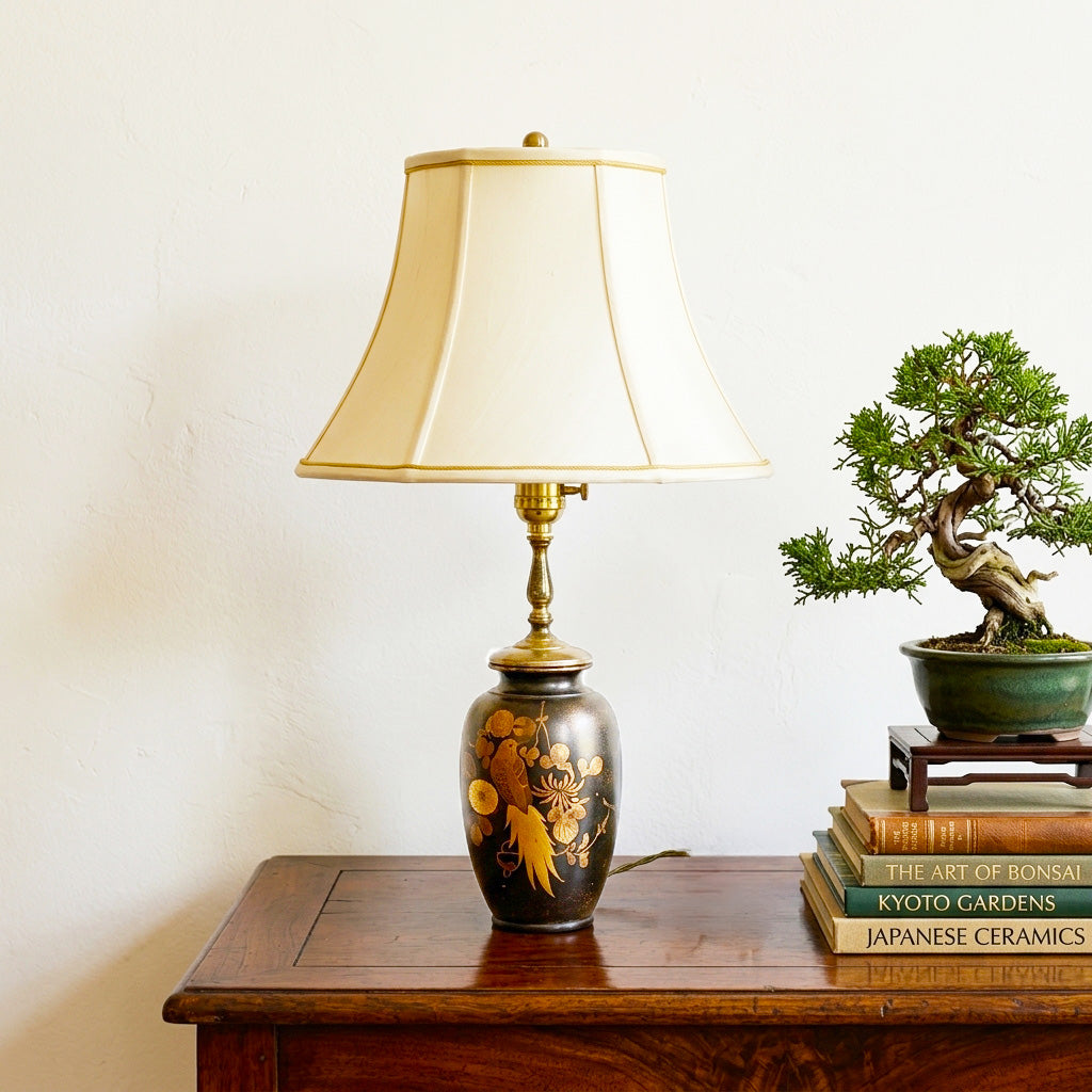 A table lamp with a cream-colored pleated shade and a dark brown glossy ceramic base decorated with gold floral and bird motifs, styled on a wooden side table next to a small potted bonsai tree and books.