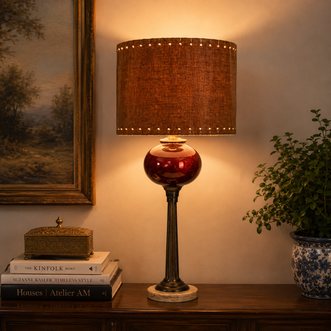 A table lamp with a brown textured drum shade, a glossy red glass globe base, and a dark metal stem, lit warmly on a wooden console table in a styled room setting.